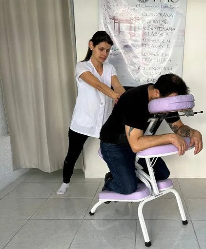 Uma profissional de saúde, aparentemente uma massoterapeuta, realiza um tratamento de massagem em uma pessoa deitada em uma cadeira de massagem.