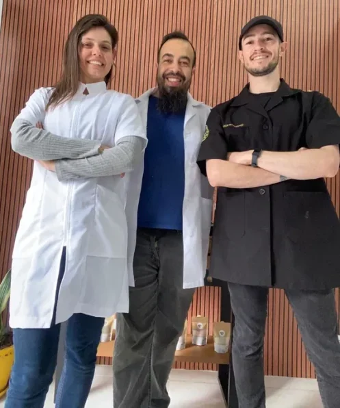Foto de três profissionais de massoterapia posando juntos em frente a uma parede de madeira.
