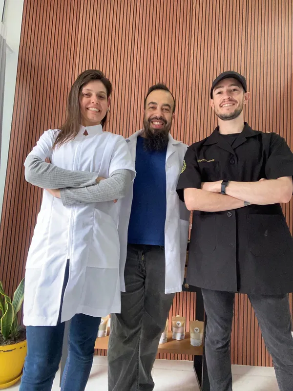 Foto de três profissionais de massoterapia posando juntos em frente a uma parede de madeira.