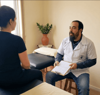 Quem Não Pode Fazer Massoterapia? Contraindicações Que Importam Um massoterapeuta do Espaço TAO com um jaleco branco e prancheta conversa com uma paciente sentada em uma maca de massagem em um consultório bem iluminado. O massoterapeuta explica as contraindicações da massoterapia durante a anamnese.