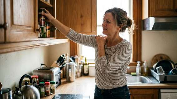 Dor no ombro ao levantar o braço: o que costuma estar por trás (e o que ajuda) Dor no ombro: Mulher com expressão de dor a segurar o ombro direito, enquanto tenta alcançar um objeto numa prateleira alta de armário de cozinha.
