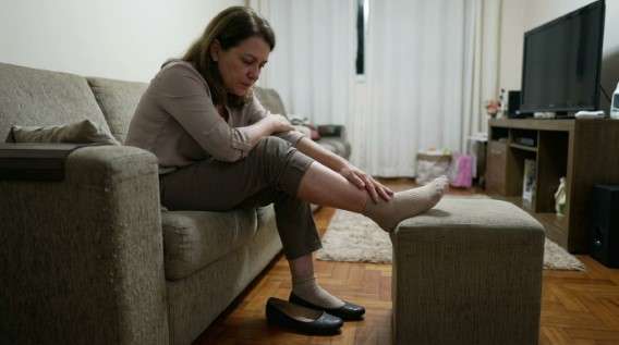 O dia inteiro a trabalhar em pé: o que isso faz com a sua lombar e as suas pernas Trabalhar em pé: Uma mulher sentada num sofá, com o pé apoiado num pufe, a massajar a própria perna com uma expressão de cansaço